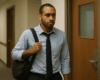 Homme en tenue de bureau debout dans un couloir d’entreprise, sac à l’épaule, regard hésitant devant une porte, songeant à l'abandon de poste pour dépression.
